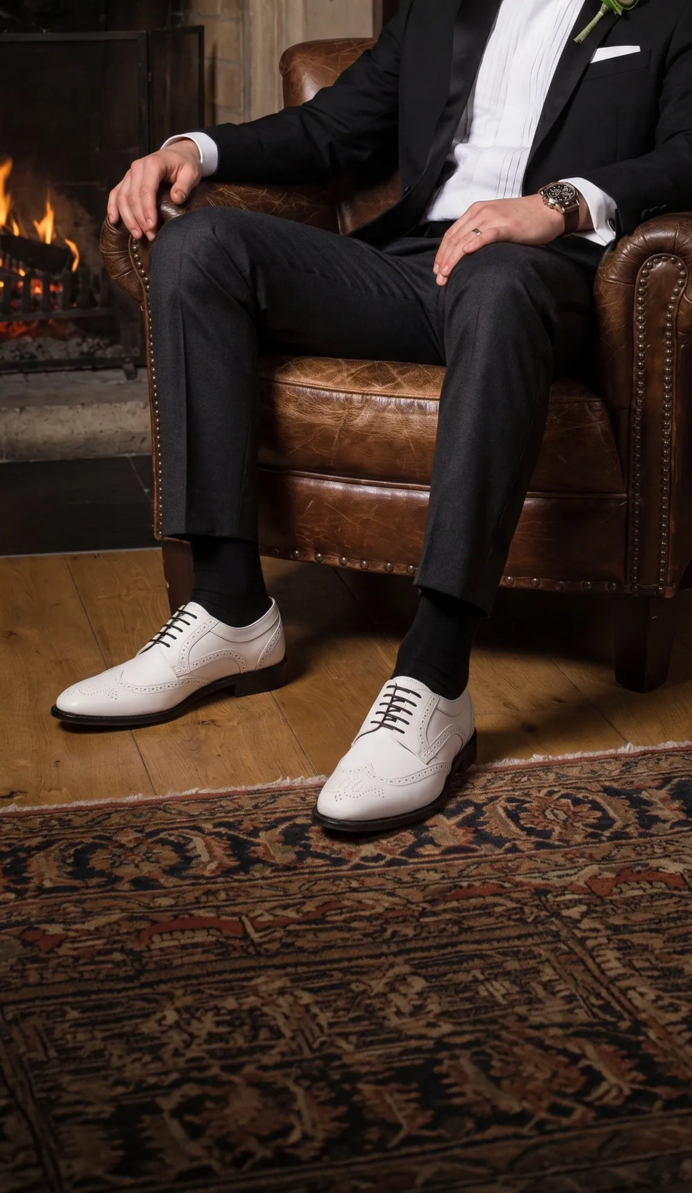 Man in a black tuxedo wearing white and black brogue shoes beside a fireplace in Mayfair after midnight